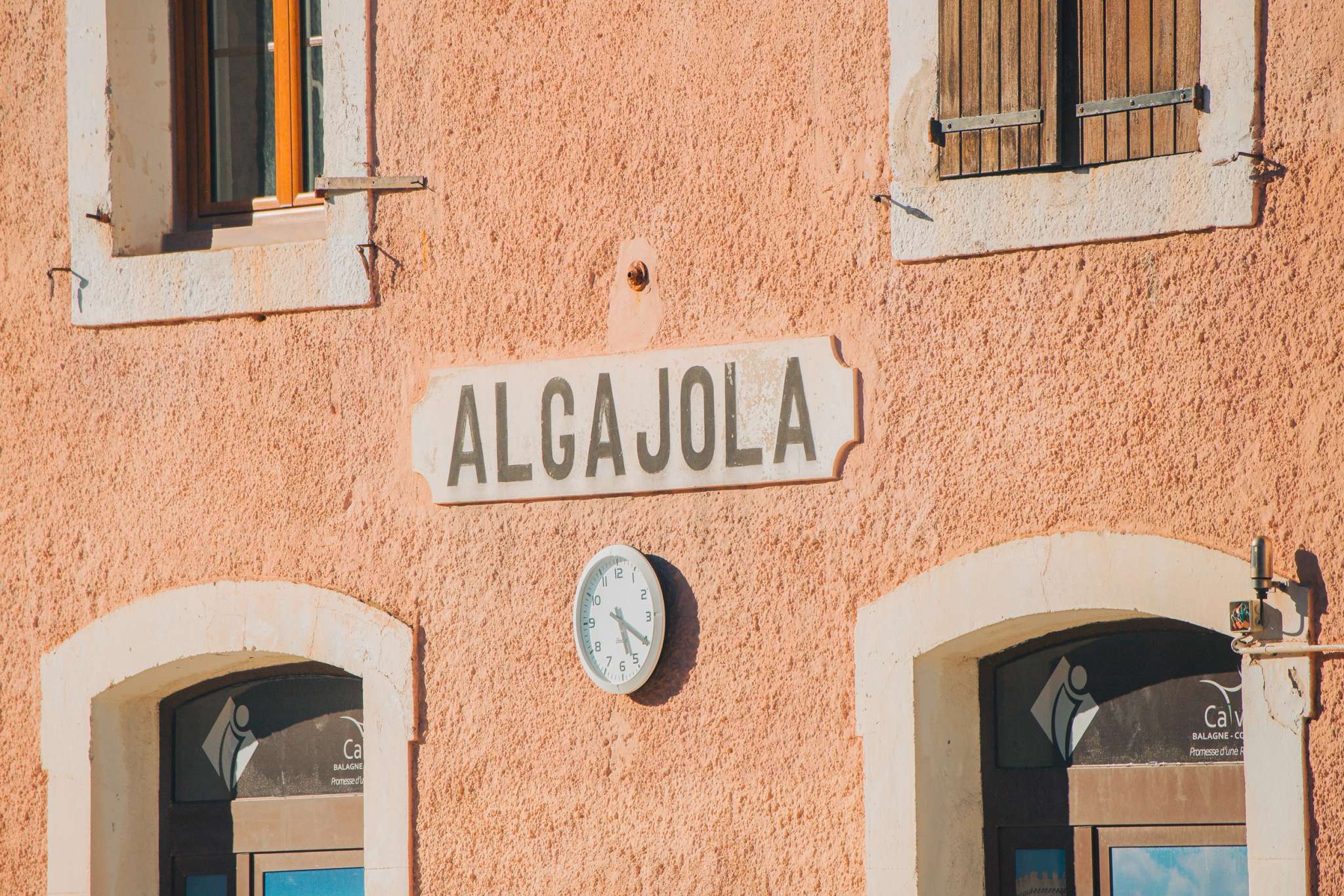 Façade d'un bâtiment en pierre ocre avec enseigne Algajola, horloge et portes voûtées en Corse